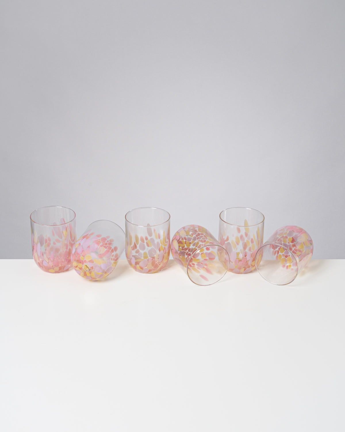 Fünf bunte, gläserne Becher mit einem rosa und gelben Muster stehen und liegen auf einem weißen Tisch.
