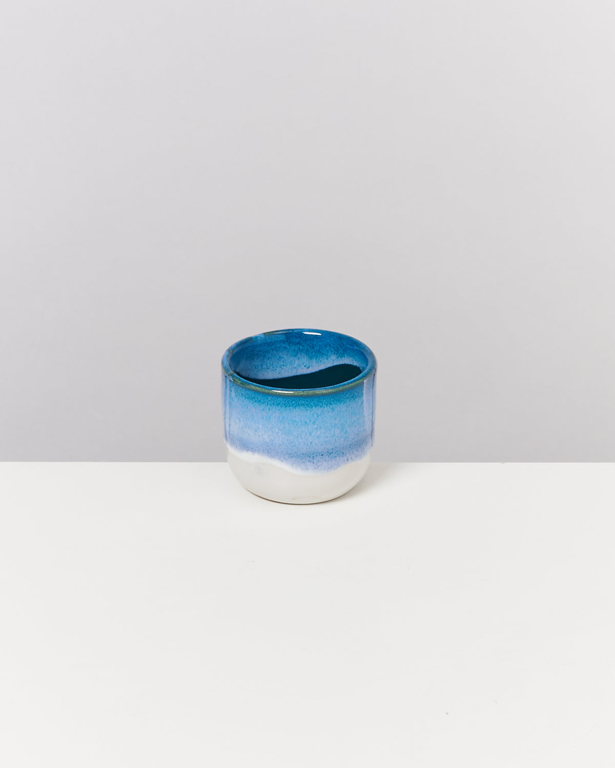 Kleiner, blauer Keramikbecher mit weißem Boden auf einer weißen Oberfläche vor grauem Hintergrund.