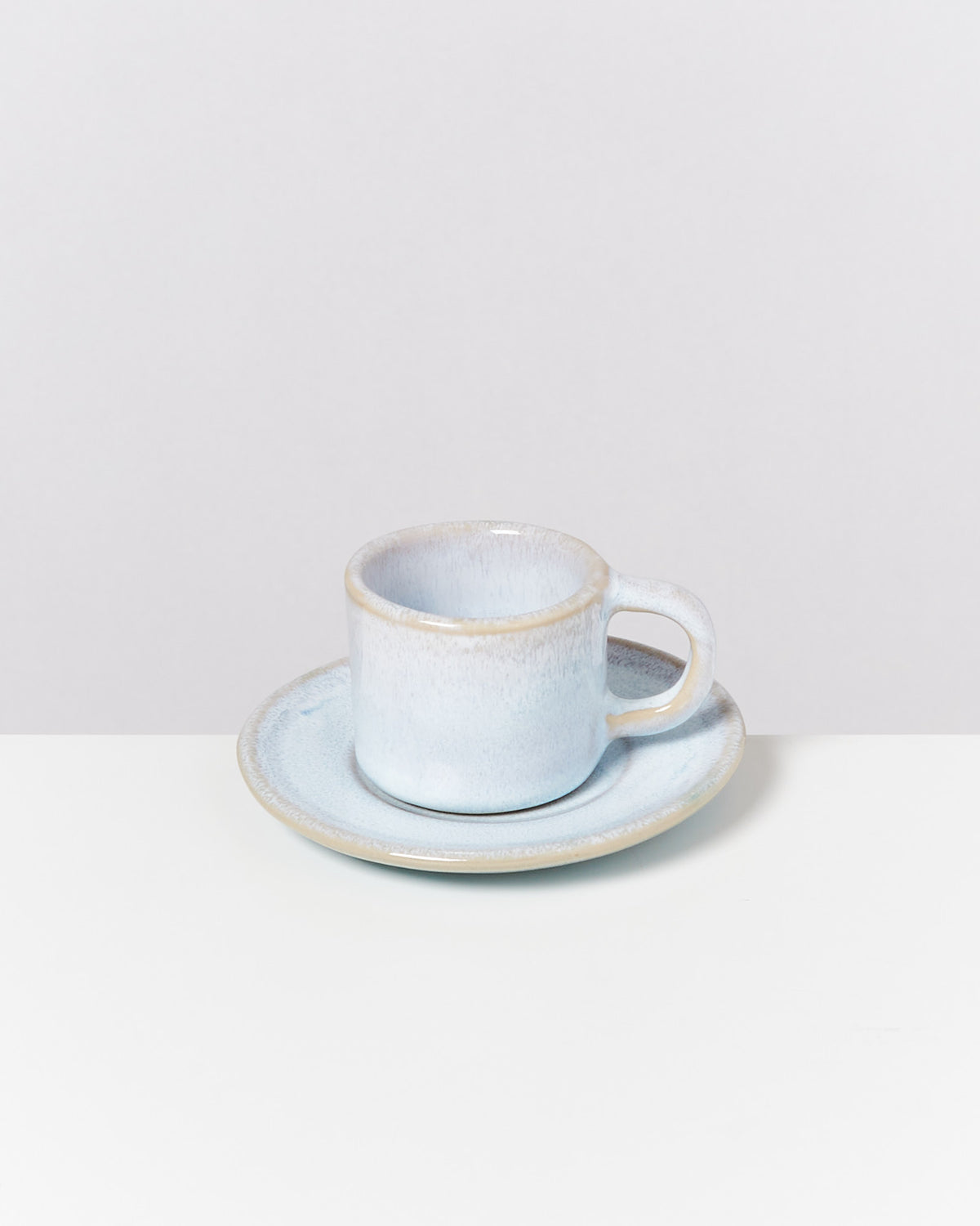 Blaue Keramiktasse auf einer passenden Untertasse, auf einem weißen Tisch vor einem hellgrauen Hintergrund.