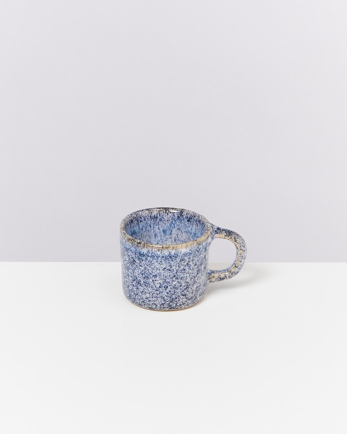 Blaue, gesprenkelte Keramiktasse mit Henkel auf einem weißen Tisch vor grauem Hintergrund.