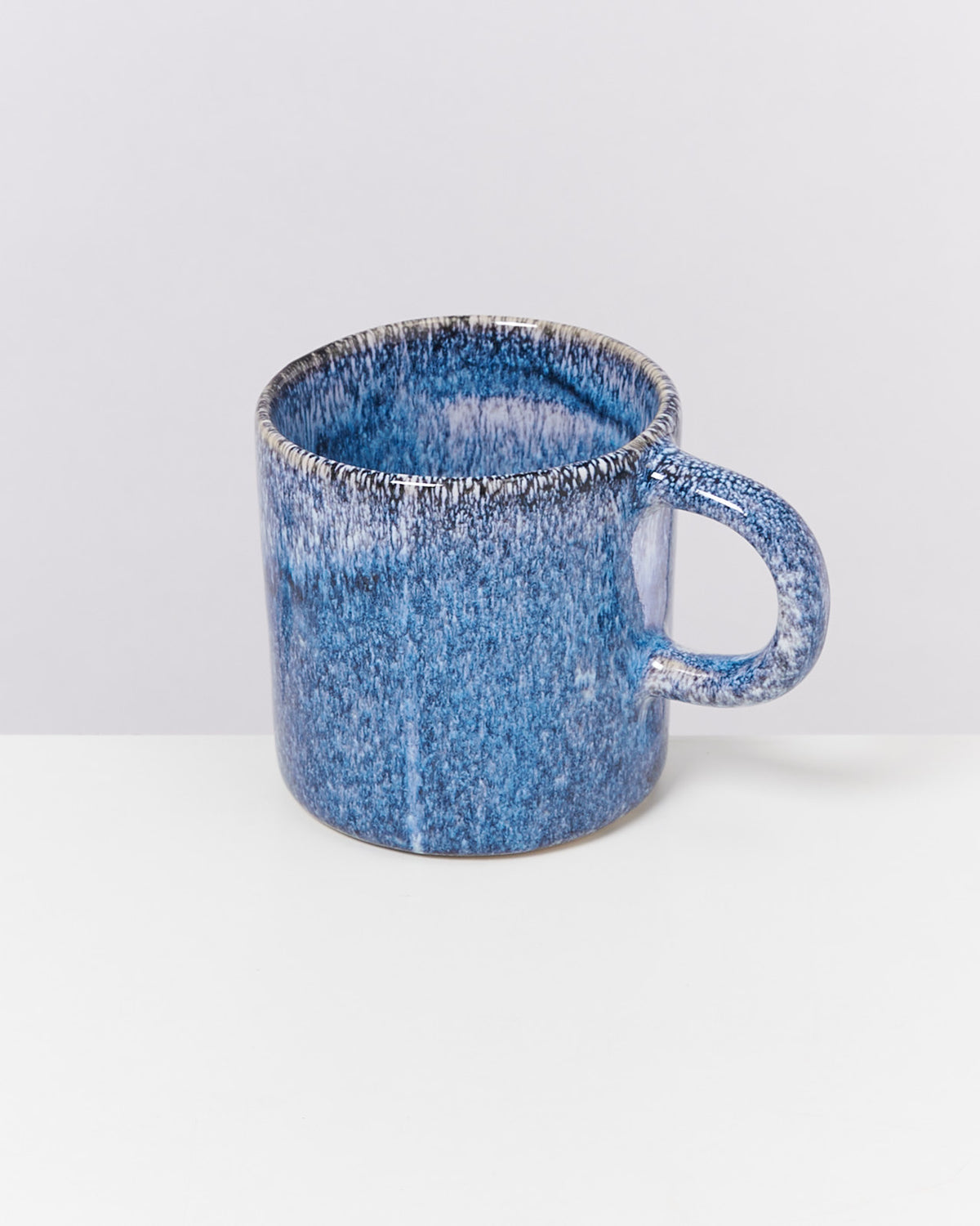 Blauer Keramikbecher mit gräulicher Maserung auf einem weißen Tisch vor einem hellen Hintergrund.