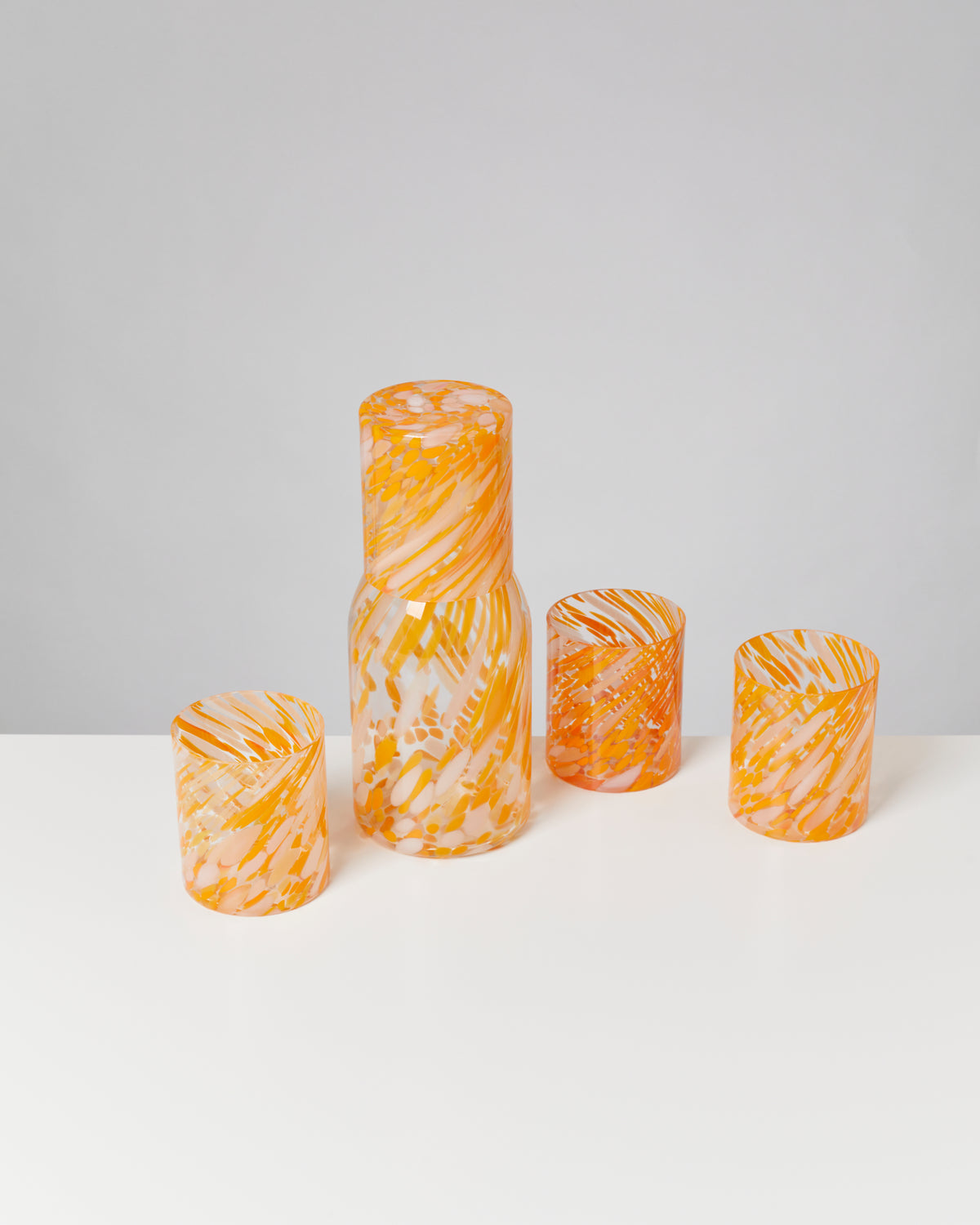 Orange gemusterte Glaskaraffe mit drei passenden Gläsern auf einem weißen Tisch.