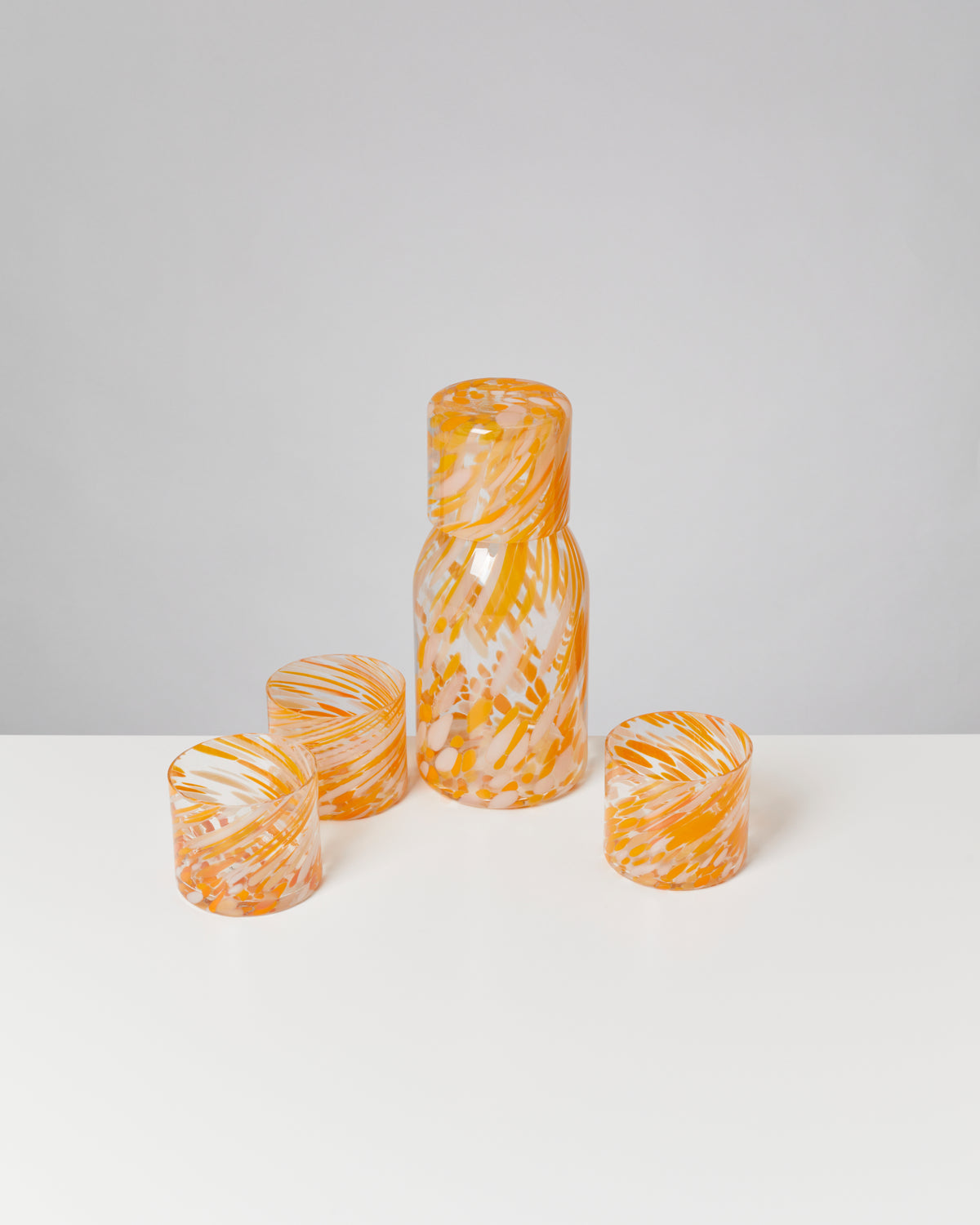 Orangene Glaskaraffe mit Deckel und drei passende Gläser mit abstraktem Muster auf weißer Oberfläche.