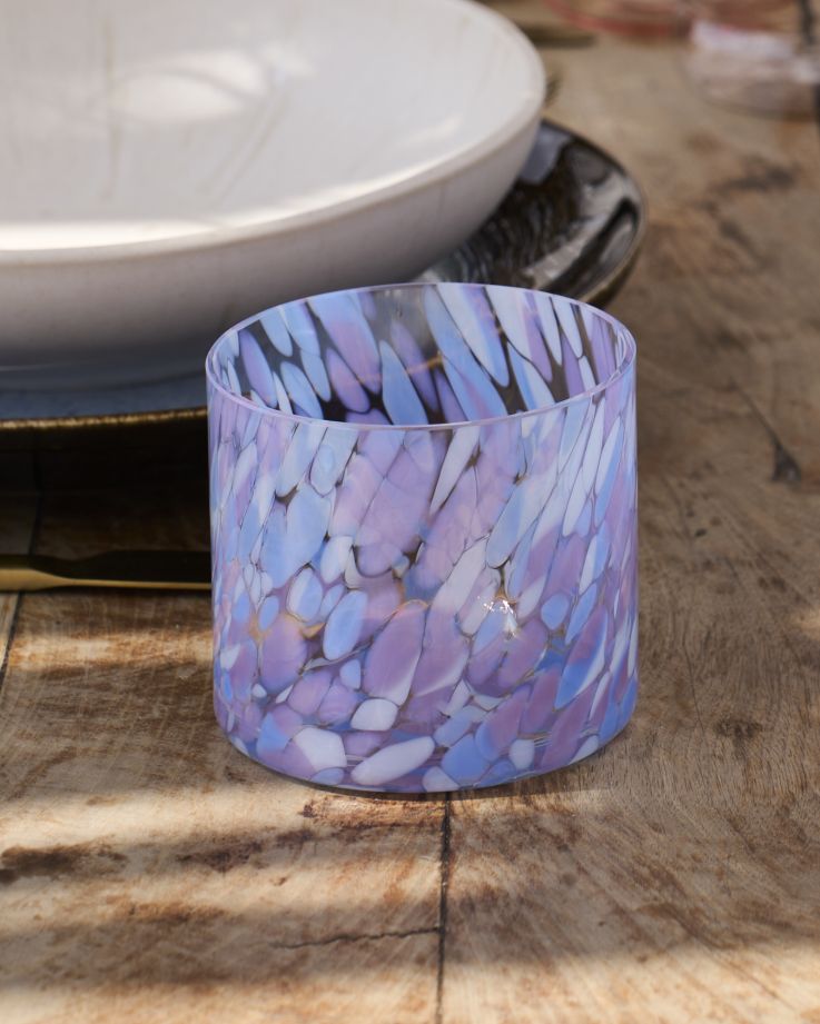 Buntes Glas mit lila und blauen Mustern auf einem Holztisch, mit einem weißen Teller im Hintergrund.