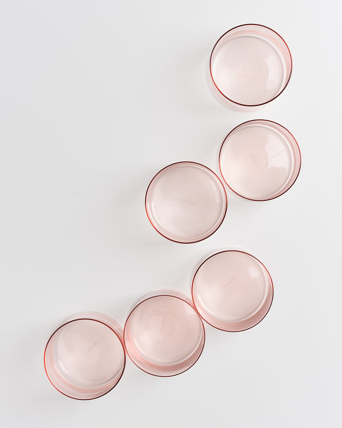 Sechs rosa Gläser mit rotem Rand sind auf einer weißen Fläche in zwei schrägen Reihen angeordnet.