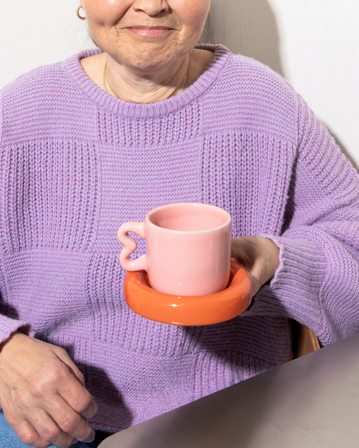 Ältere Frau in lilafarbenem Pullover hält eine rosa Tasse mit einem orangefarbenen Untersetzer in der Hand.
