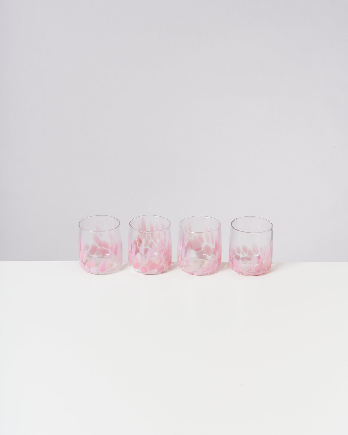 Vier Gläser mit rosa Muster stehen in einer Reihe auf einem weißen Tisch vor hellem Hintergrund.