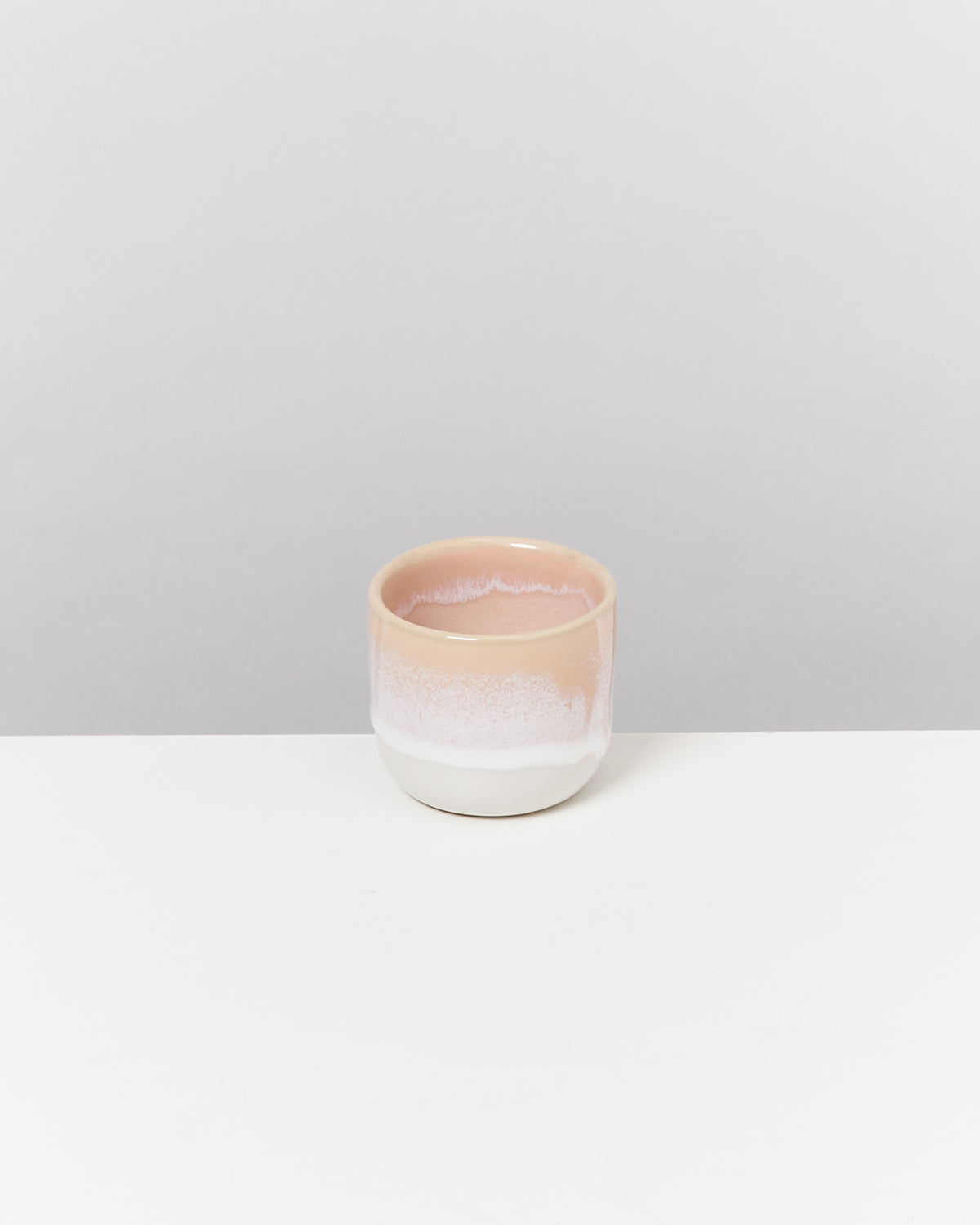 Keramikbecher mit rosa und weißem Farbverlauf steht auf weißer Oberfläche vor grauem Hintergrund.