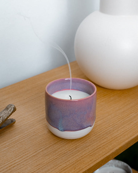 Lila Kerze in einem keramikähnlichen Kerzenhalter auf einem Holztisch, mit Rauchfaden in der Luft.