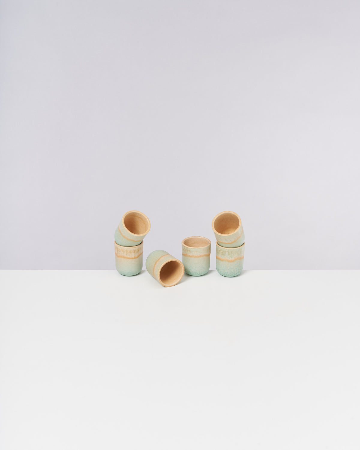 Fünf kleine Keramikbecher mit blauer und brauner Glasur auf weißem Untergrund, drei stehend und zwei liegend.