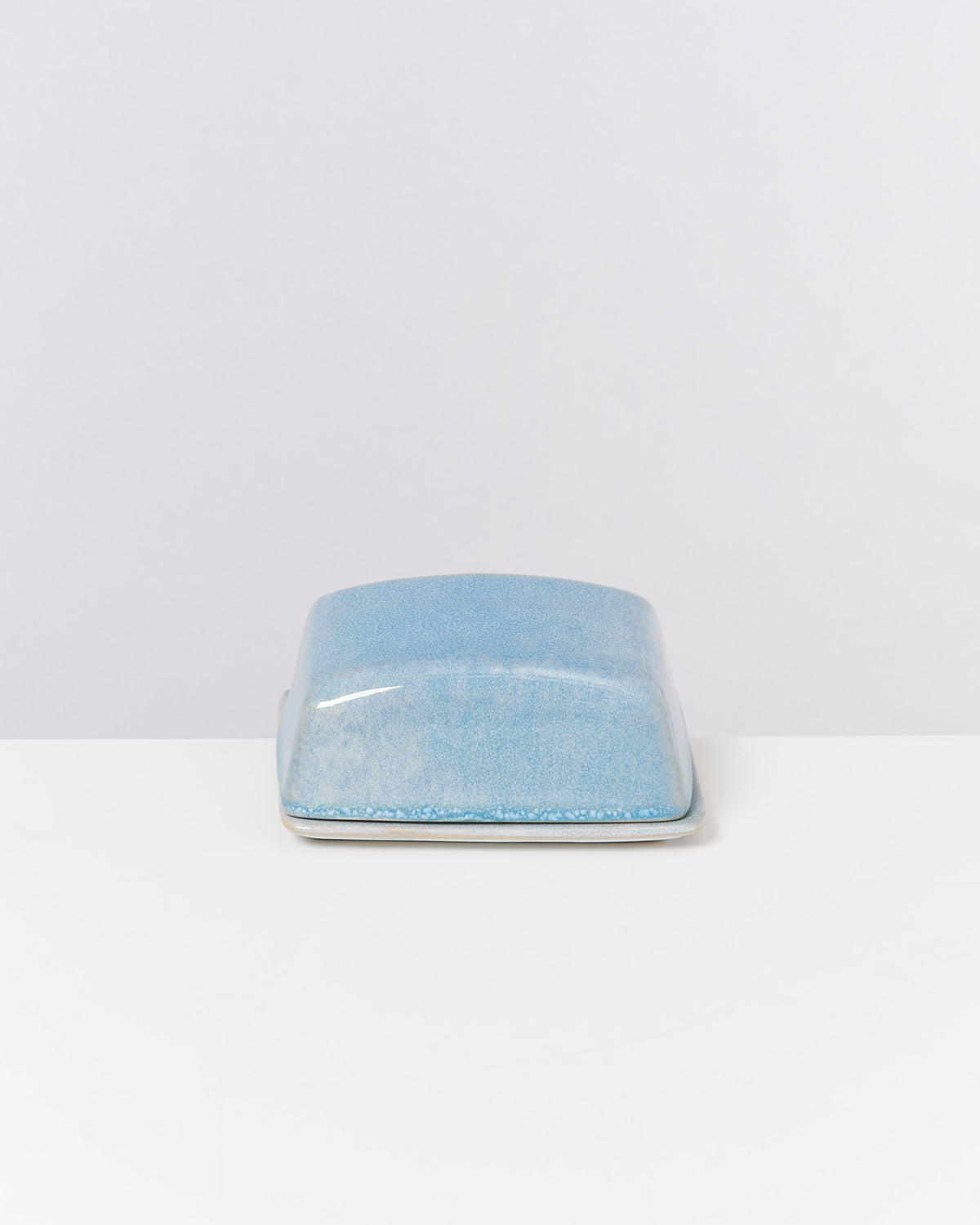 Blaue, rechteckige Keramikbutterdose mit Deckel auf einem weißen Tisch vor hellem Hintergrund.