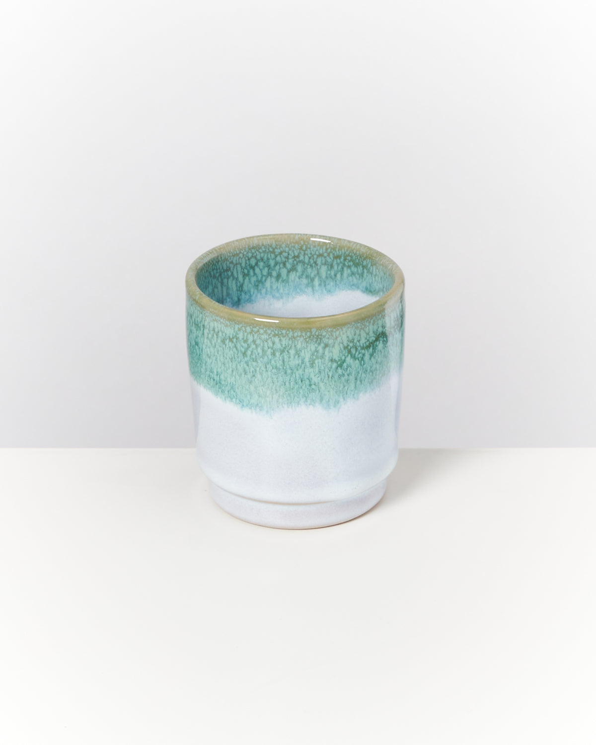 Keramikbecher mit graugrüner Glasur am oberen Rand steht auf einem weißen Hintergrund.