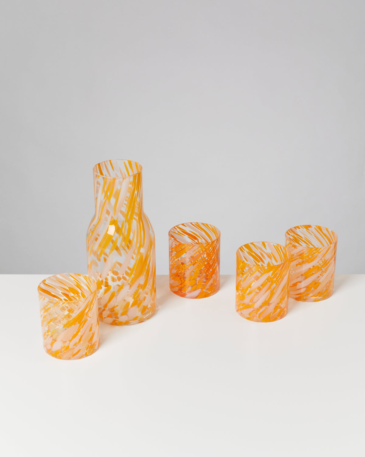 Fünf orange-weiß gemusterte Trinkgläser stehen auf einem weißen Tisch vor grauem Hintergrund.