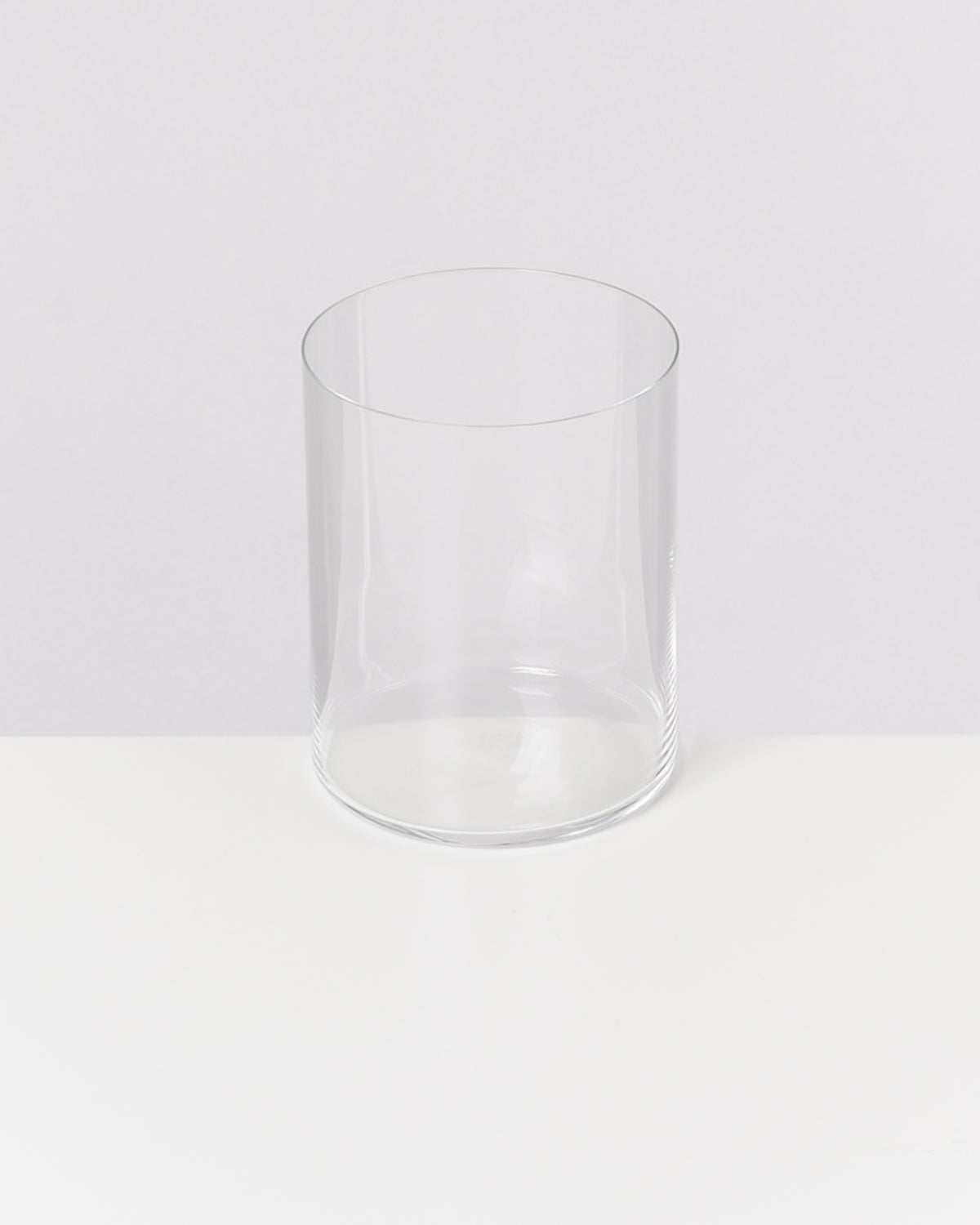 Ein leeres, transparentes Trinkglas steht auf einem weißen Tisch vor einem grauen Hintergrund.