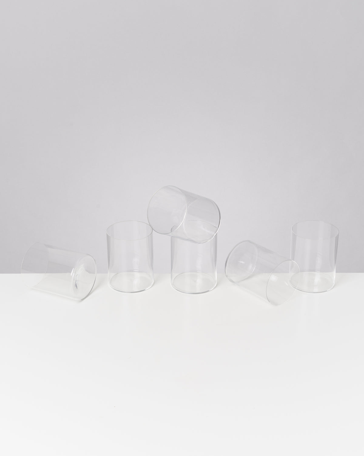 Fünf leere, transparente Gläser liegen und stehen auf einem weißen Tisch vor grauem Hintergrund.