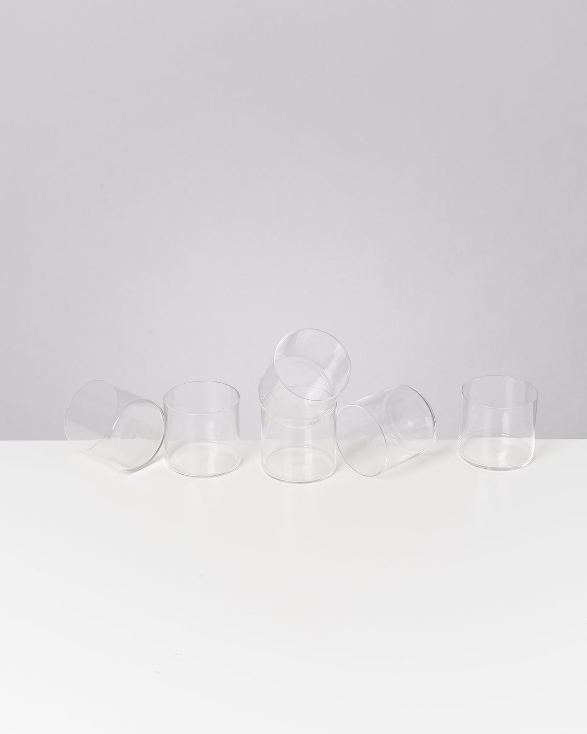 Sechs umgekippte, transparente Gläser auf einem weißen Tisch vor grauem Hintergrund.