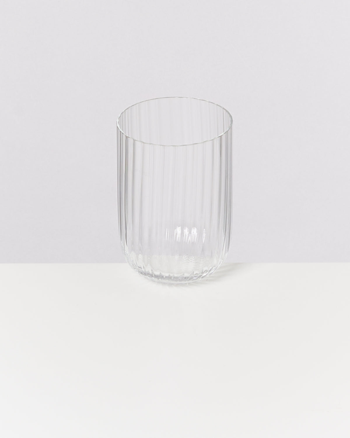 Ein geripptes, klares Glas steht auf einer weißen Oberfläche vor einem grauen Hintergrund.