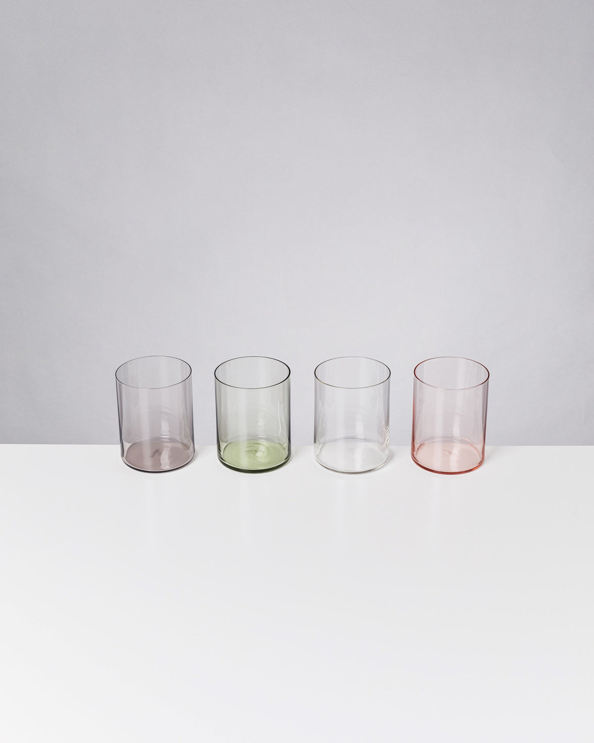 Vier transparente, farbige Gläser in Lila, Grün, Klar und Rosa auf einem weißen Tisch vor grauem Hintergrund.