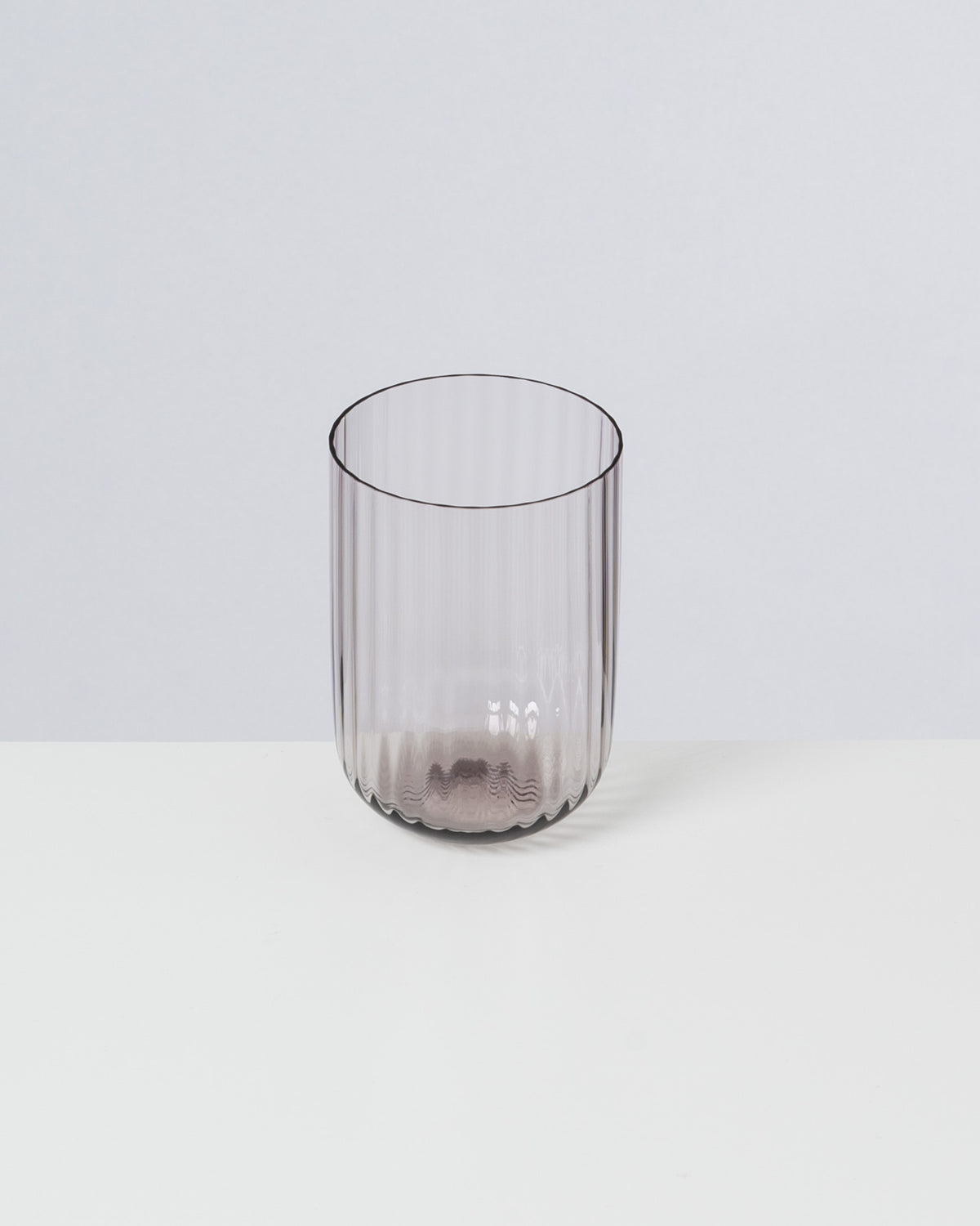 Ein durchsichtiges, geripptes Trinkglas steht auf einem weißen Tisch vor einem grauen Hintergrund.