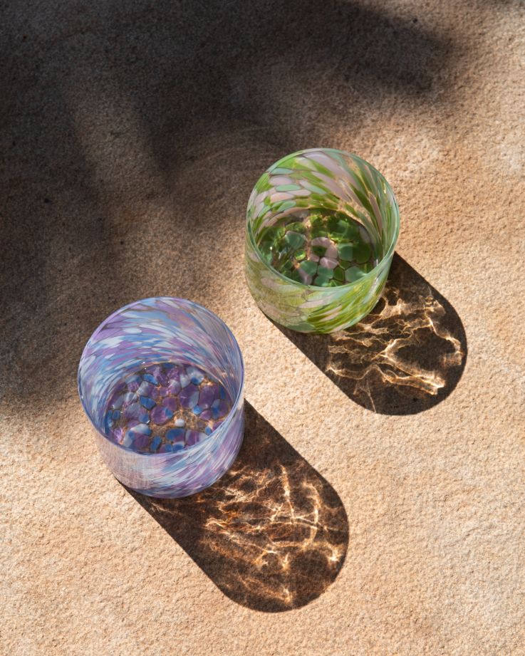 Zwei bunte Gläser auf einem sonnenbeschienenen Sandboden werfen lange, farbige Schatten.