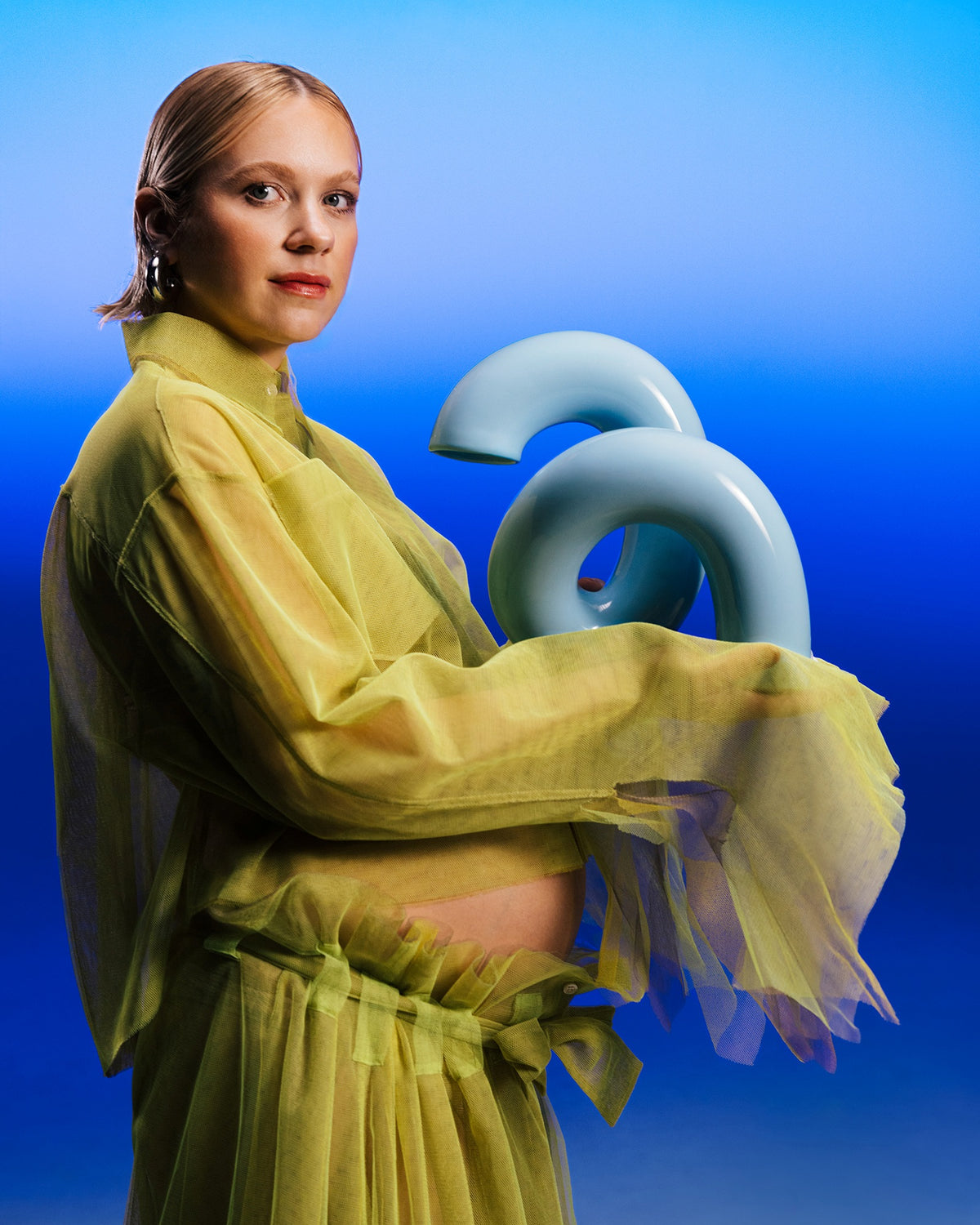 Eine Frau in einem gelben Kleid hält eine blaue Skulptur vor einem blau verlaufenden Hintergrund.