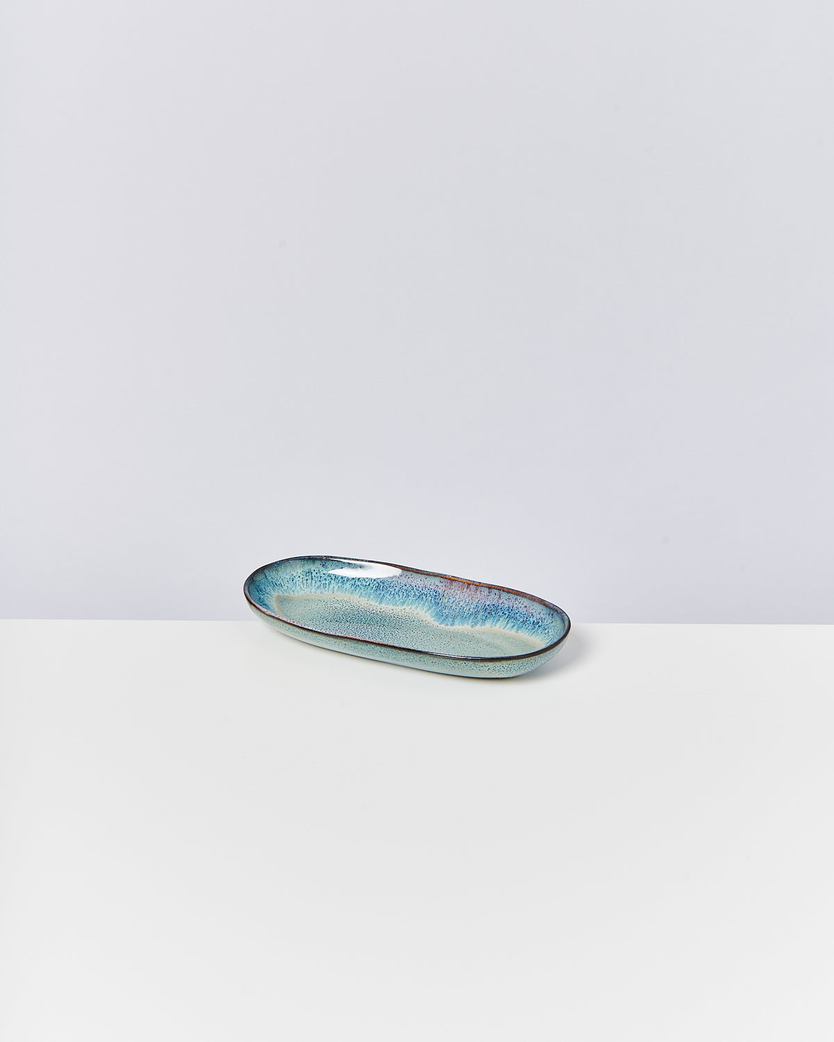 Langes, ovales Keramikschälchen in Blau- und Grüntönen auf einem weißen Tisch vor neutralem Hintergrund.