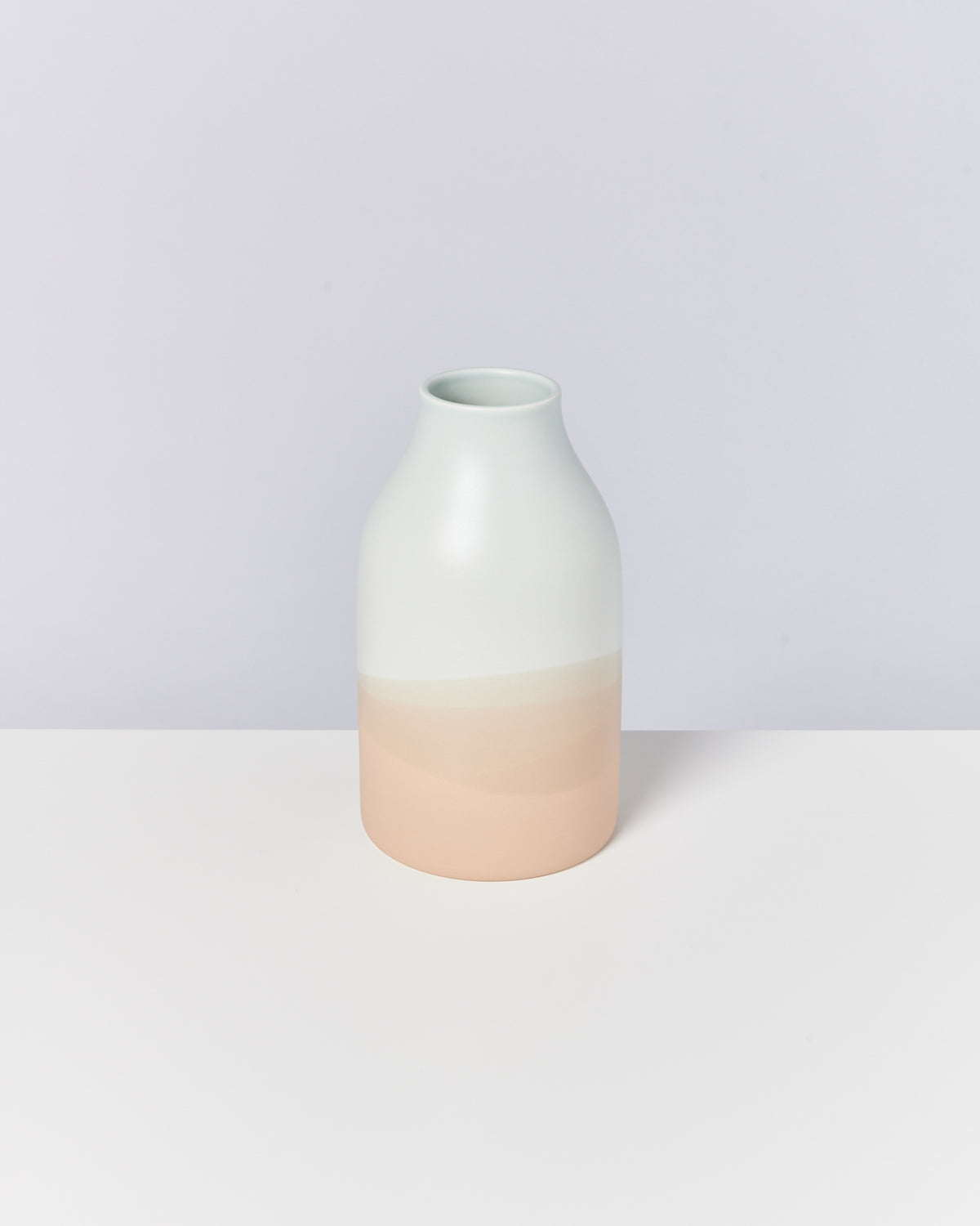 Minimalistische Vase in mattem Farbverlauf von Weiß nach Hellrosa, auf einem hellen Hintergrund stehend.