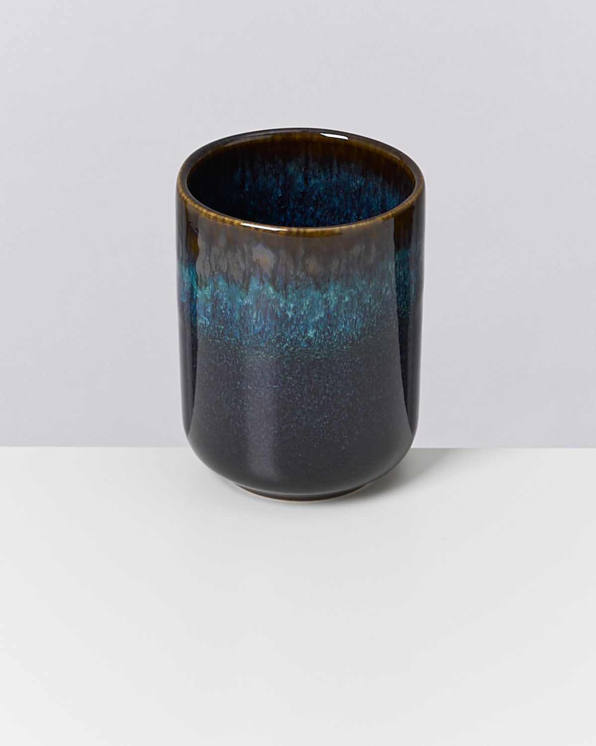 Keramikbecher mit blau-grüner Glasur und braunem Rand auf weißem Hintergrund.
