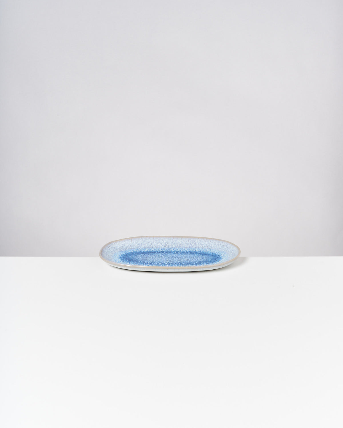 Ovale Keramikplatte mit blauer Mitte auf weißem Tisch vor grauem Hintergrund.