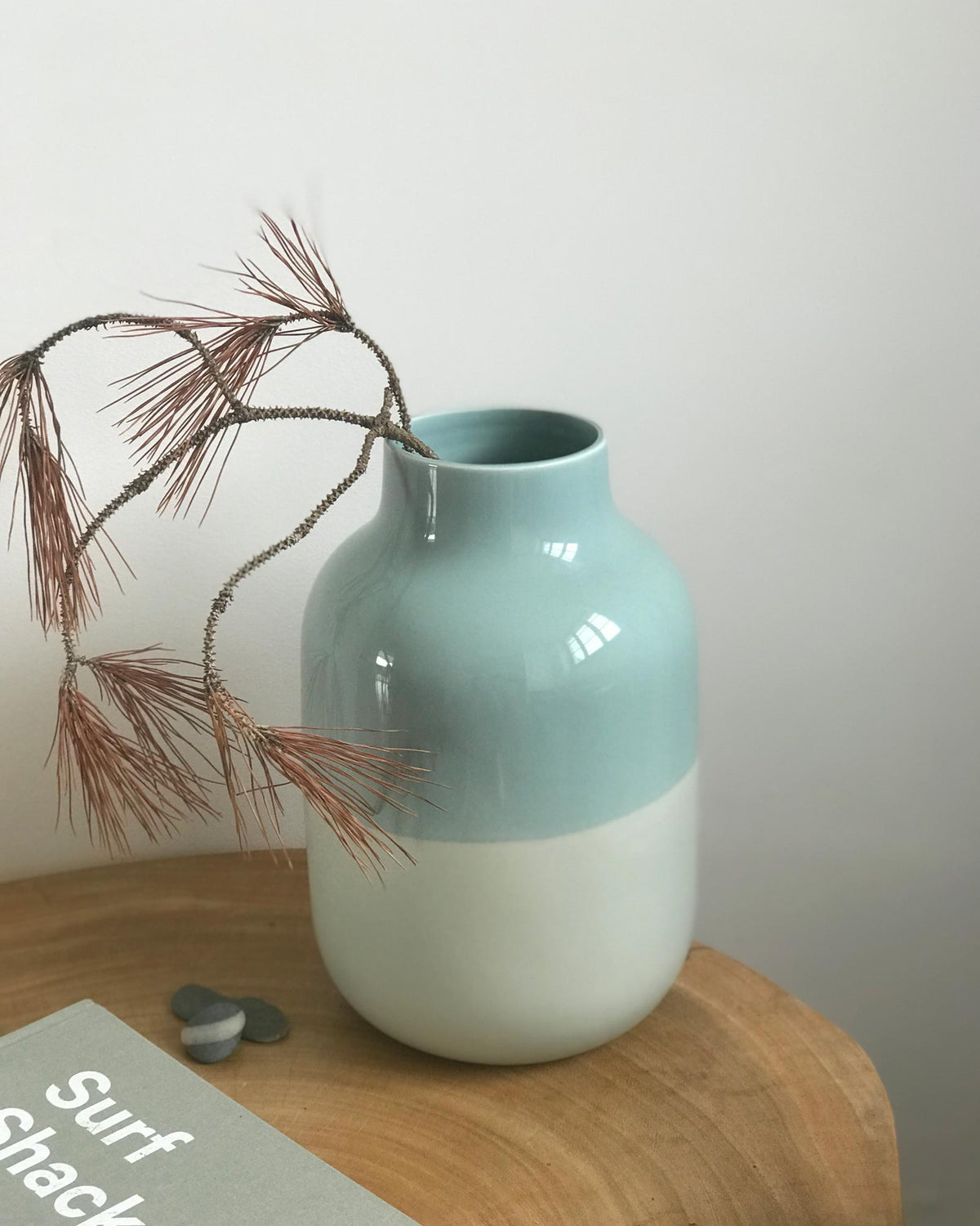 Blaue Vase mit trockenen Zweigen steht auf einem Holzbrett neben kleinen Steinen und einem Buch mit der Aufschrift „Surf Shack“.