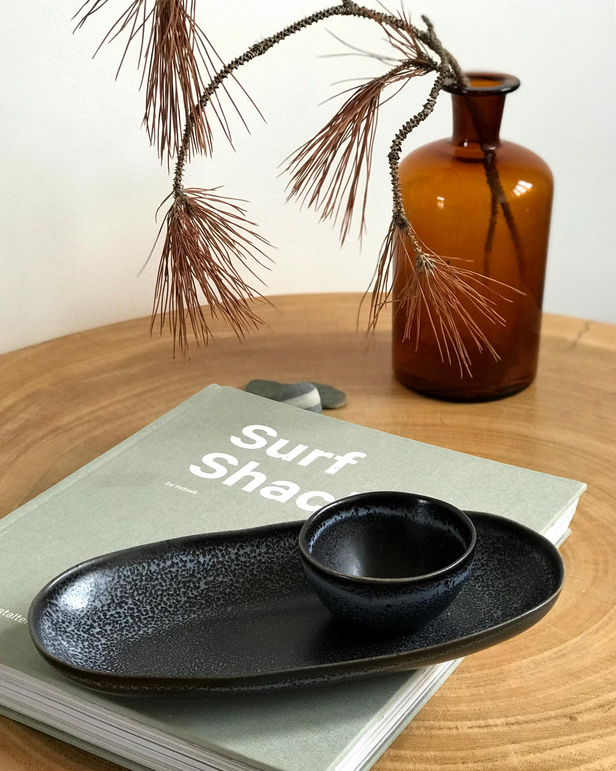 Buch mit Titel "Surf Shacks", eine braune Vase mit Zweig und schwarzes Geschirr auf einem Holztisch.