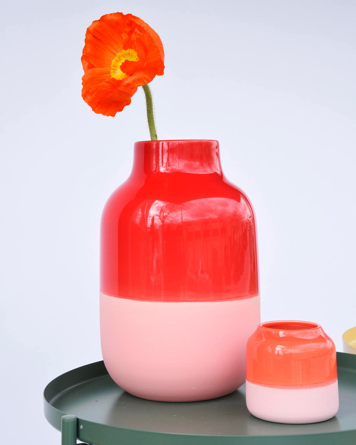 Zwei bunte Vasen, eine große und eine kleine, stehen auf einem Tisch, wobei die große Vase eine orange Blume enthält.