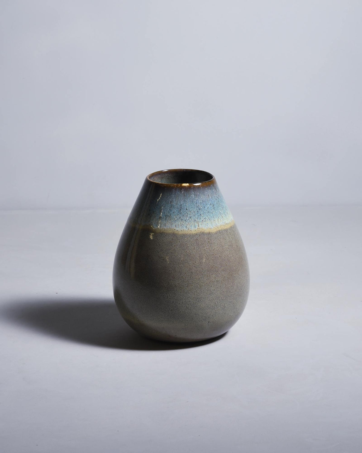 Graue Keramikvase mit blauer und brauner Glasur auf hellem Hintergrund, wirft einen Schatten nach links.