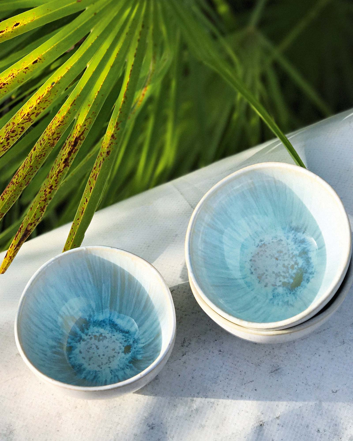 Zwei handgemachte Schalen mit blauem Muster stehen auf einem Tisch, neben ihnen grüne Palmblätter im Sonnenlicht.