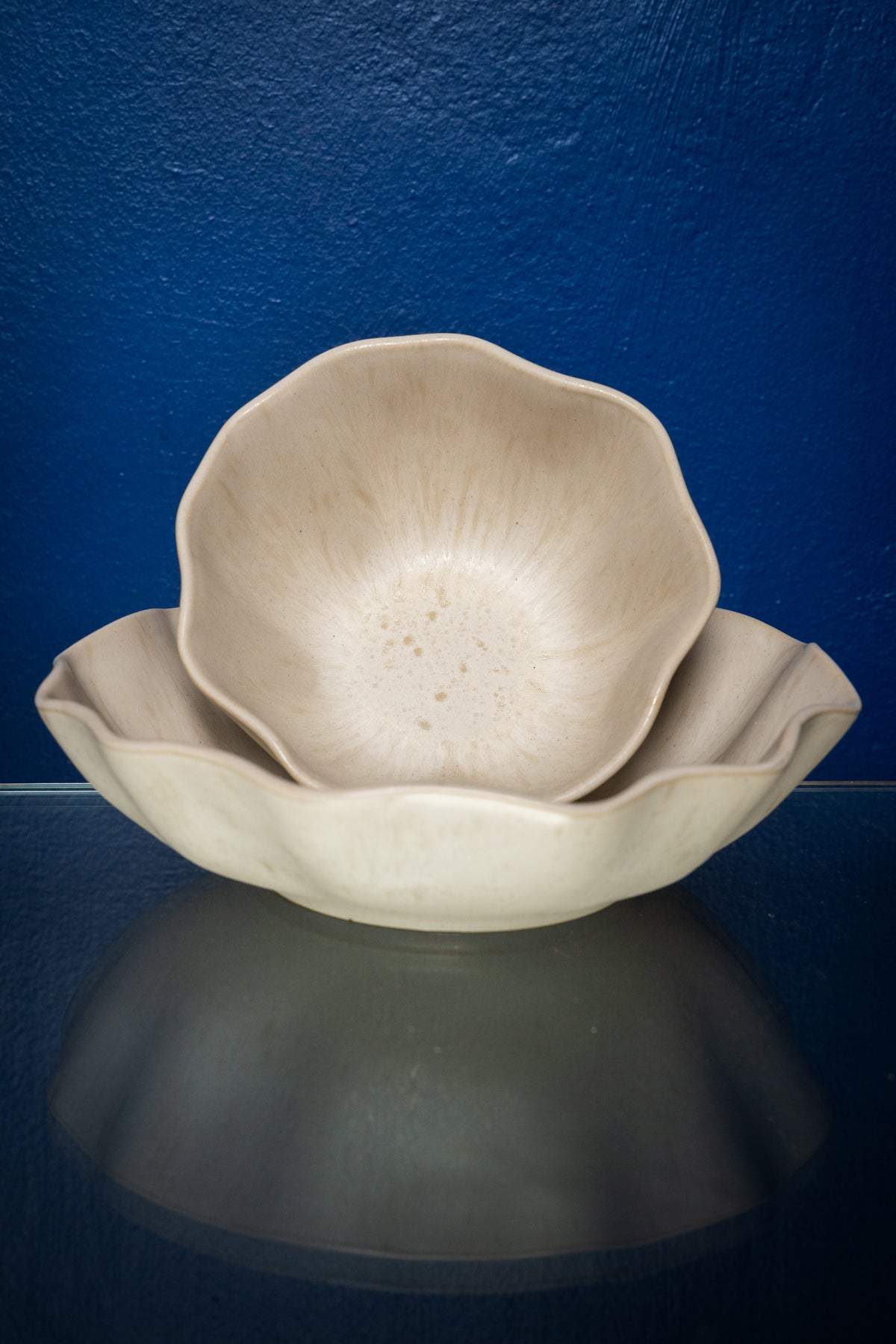 Zwei beige, blütenförmige Schalen stehen ineinander vor einer blauen Wand auf einem Glasoberfläche.