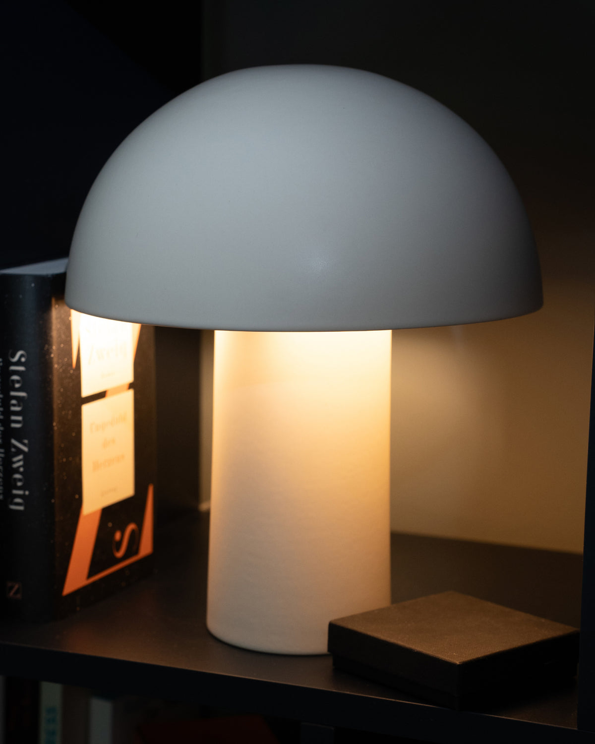 Moderne Tischlampe mit rundem Schirm, leuchtet warmes Licht aus, neben Büchern auf einem Regal platziert.