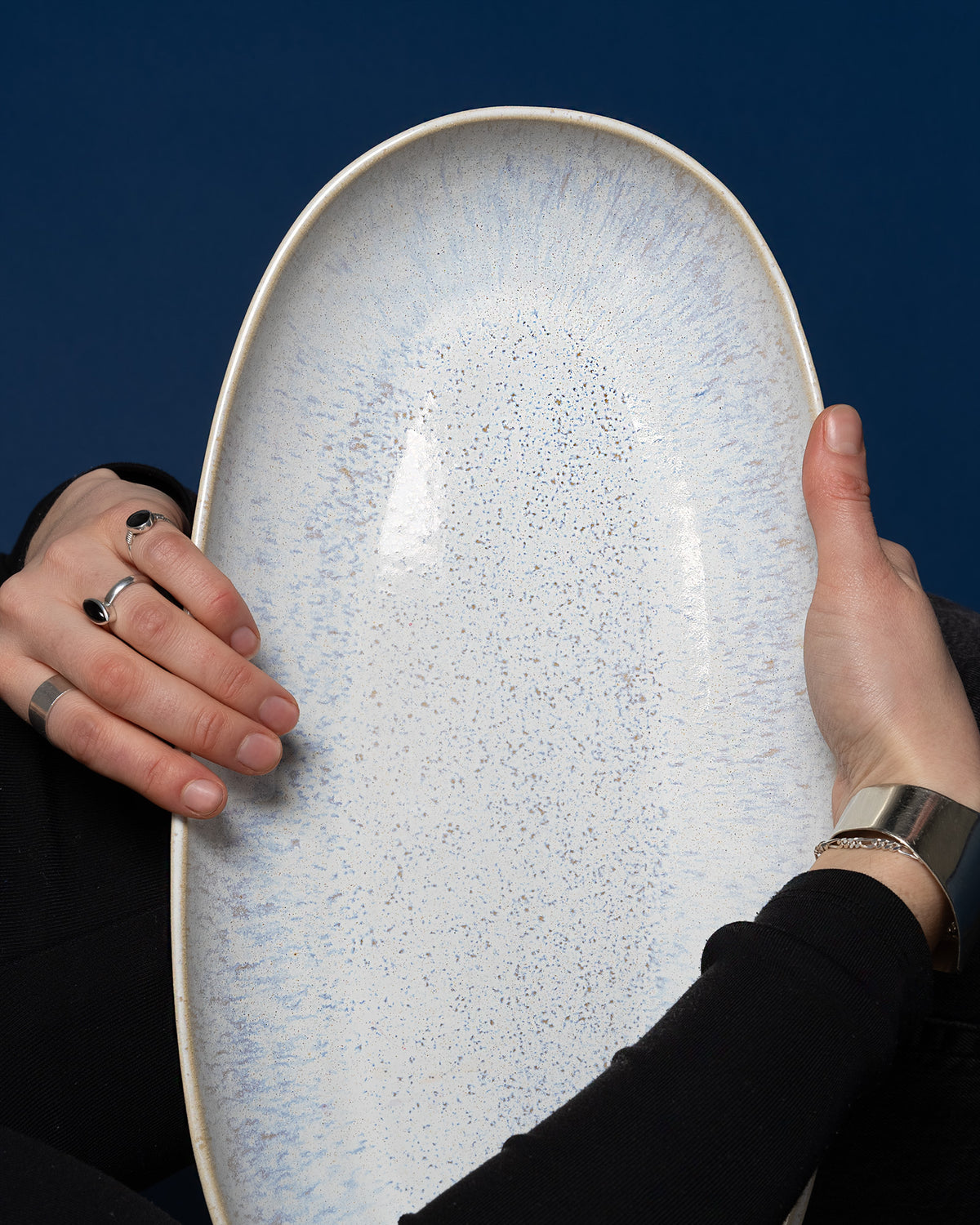 Zwei Hände halten ein ovales, weiß-blau gesprenkeltes Keramiktablett vor einem dunkelblauen Hintergrund.